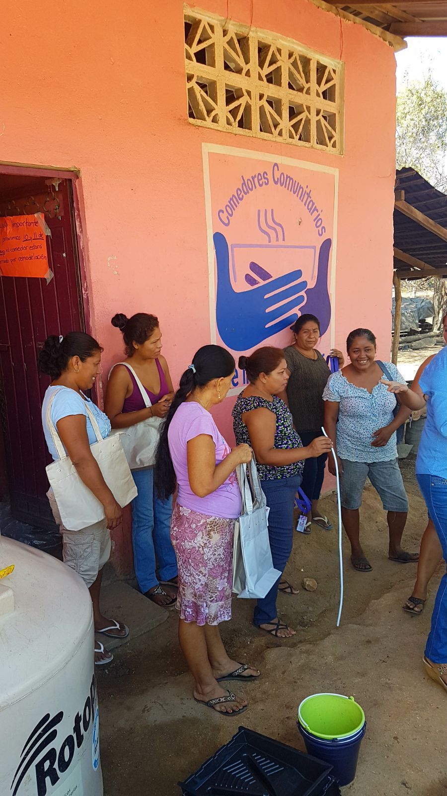 People gathered outside a pink building, standing near a wall sign and a bucket in a shaded courtyard.
