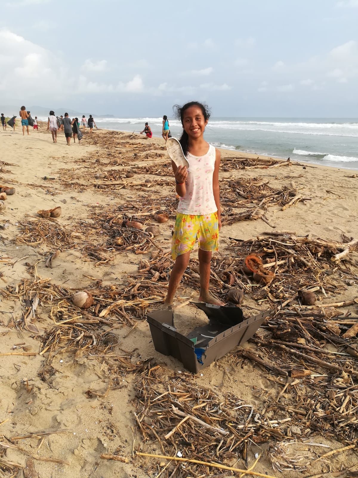 Person standing in a black container on a littered beach, holding a shovel with other people in the distance.