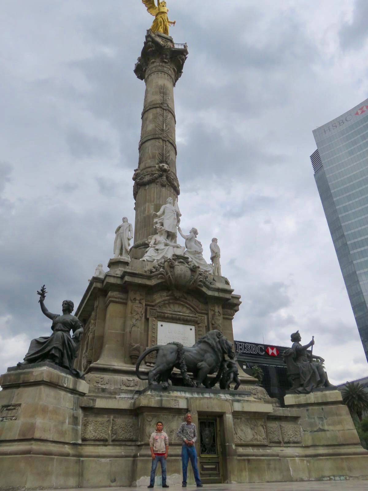 Monument with statues in a city square under a cloudy sky, with two people standing at the base.