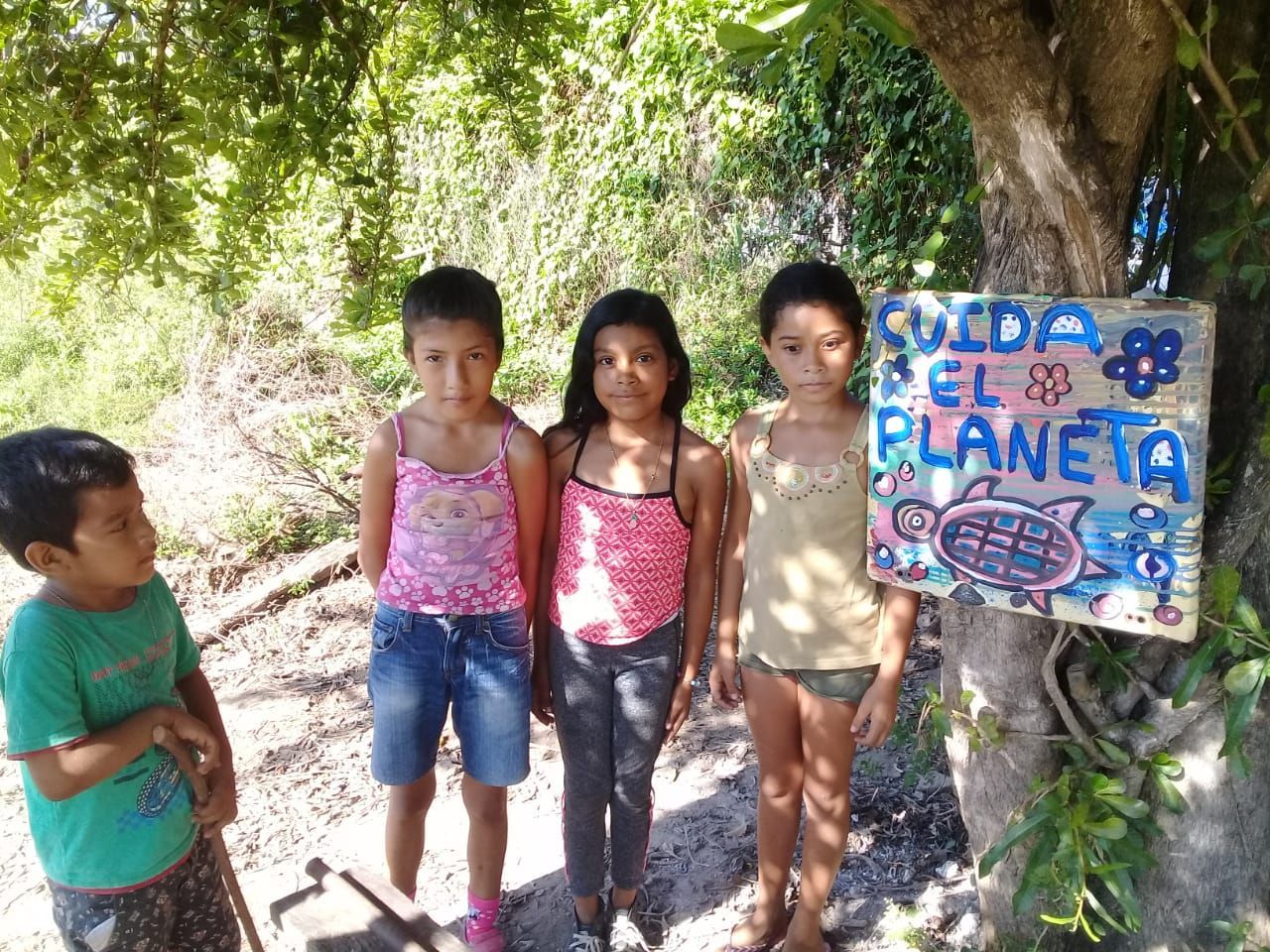 Four children stand outdoors beside a handmade sign reading “Cuida el planeta.”