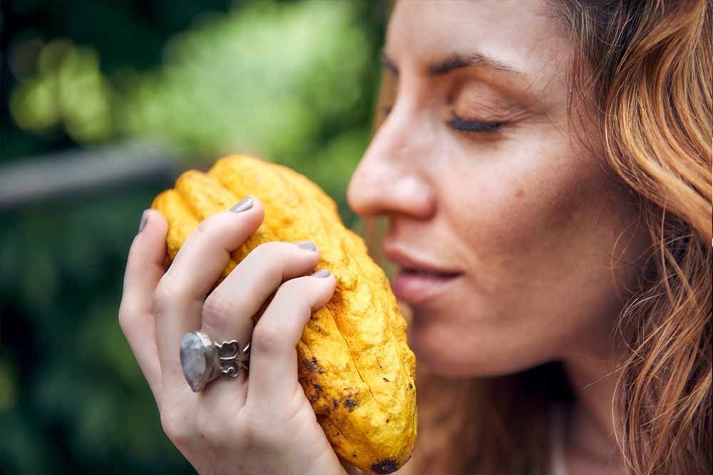 Woman smelling a yellow cacao pod, with a ring on her hand, outdoors against a blurred green background