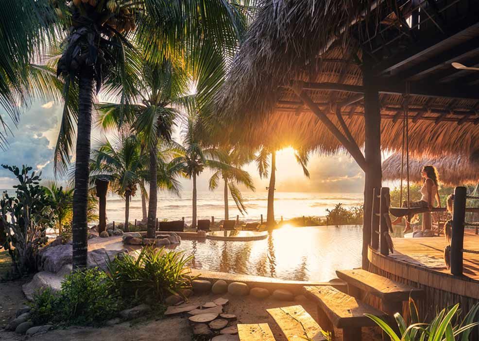 Sunset over a tropical resort pool with palm trees and thatched cabanas by the water