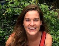 Smiling woman with brown hair in a red top, standing in front of leafy greenery