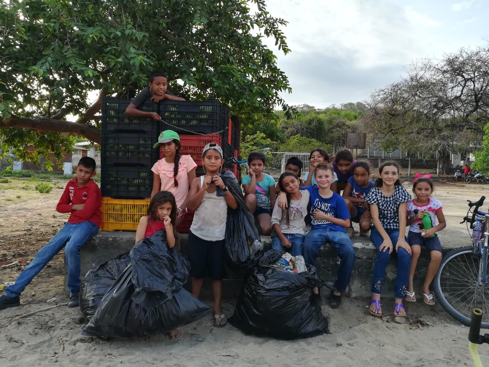 Group of children sitting outdoors by large trash bags, posing together in a park or schoolyard.