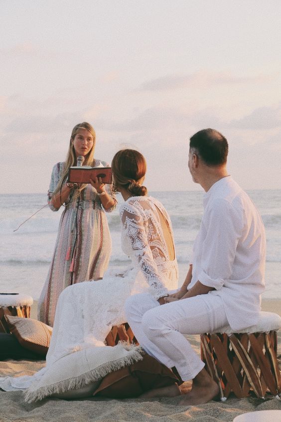 Three people in white outfits by the sea, one reading from a folder at sunset