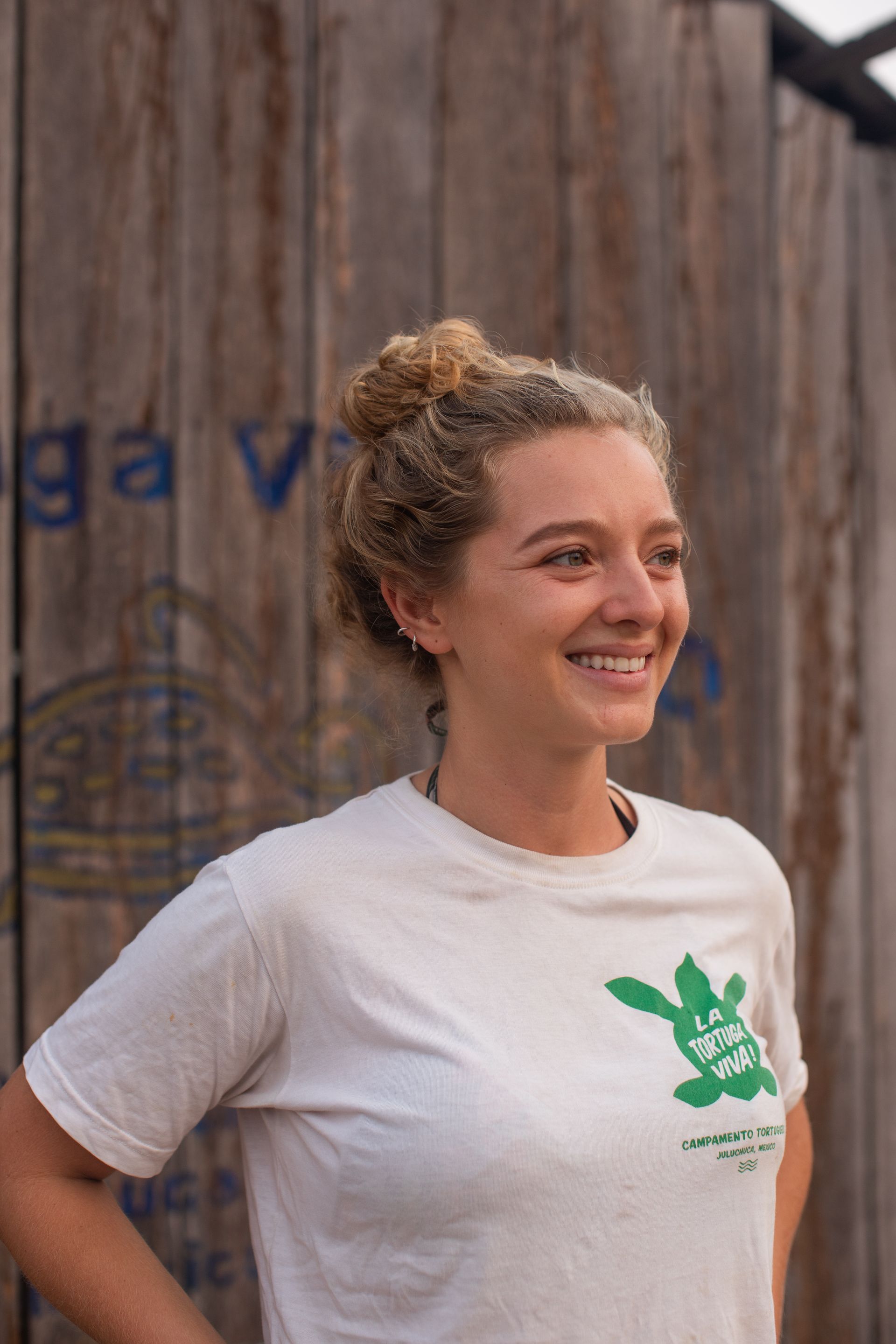 Smiling woman in a white T-shirt standing outdoors beside a wooden wall