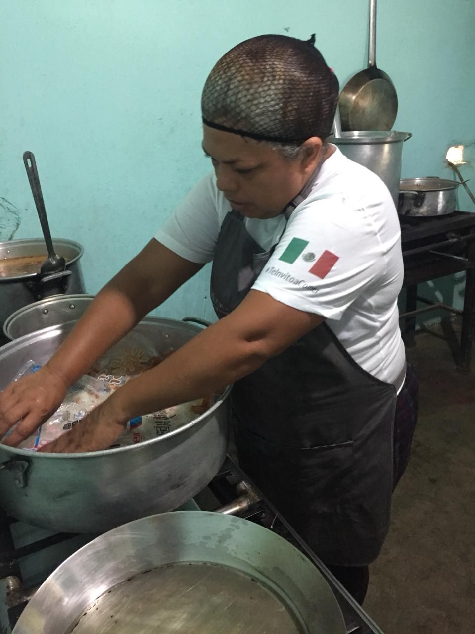Person washing food in a large metal bowl in a kitchen, wearing a white shirt and hairnet.