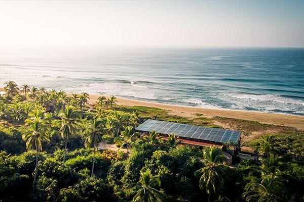 Aerial view of a tropical beachfront house surrounded by palm trees beside the ocean at sunrise.