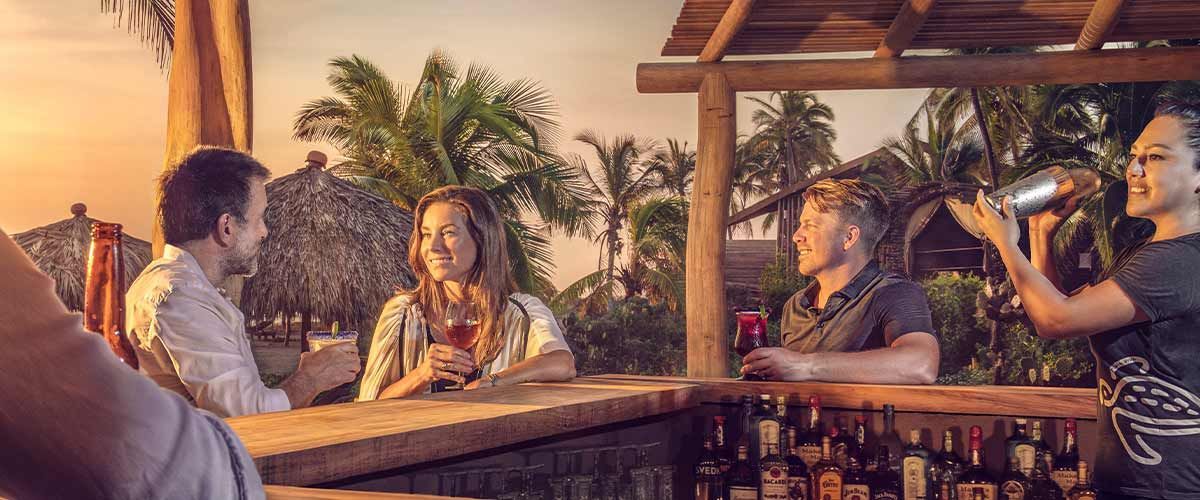 People chatting at a rooftop bar at sunset, with drinks and palm trees in the background