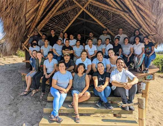 Group seated on wooden steps inside a thatched shelter, smiling for a group photo
