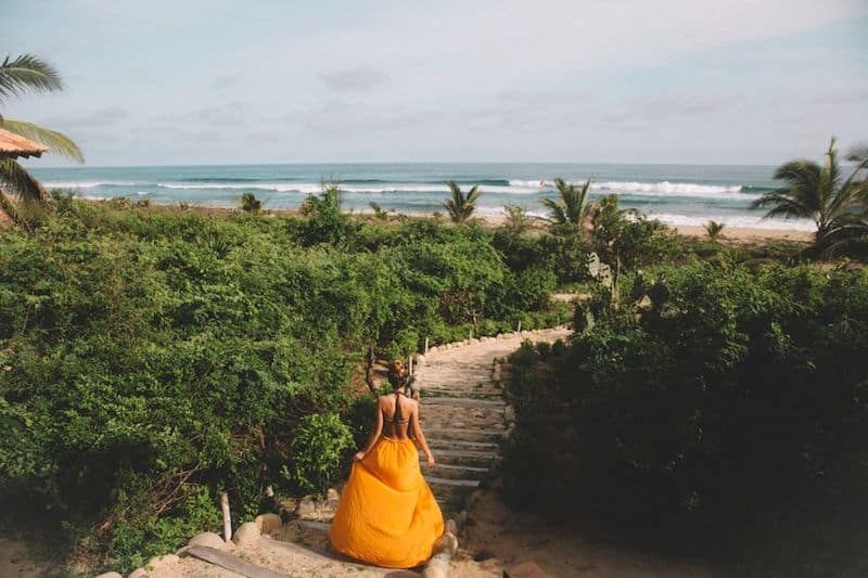 Person in a bright orange dress sits on steps overlooking a lush tropical beach and ocean