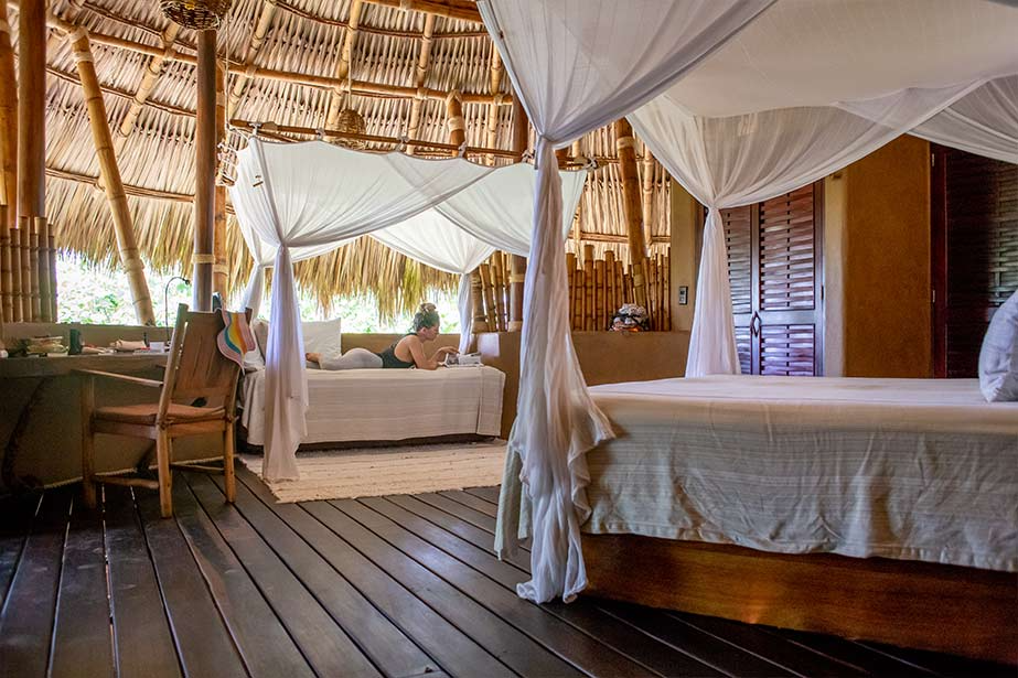 A rustic hotel room with a thatched roof, wooden floor, two canopy beds, and a person relaxing on one of the beds.