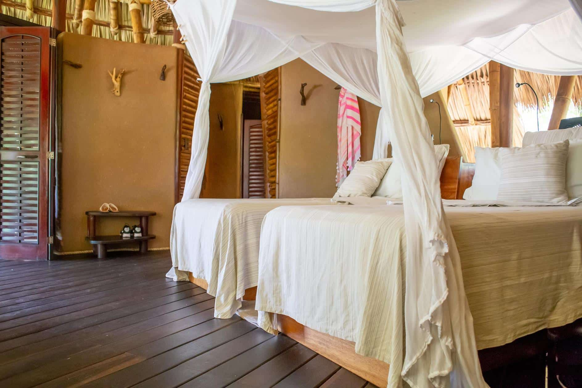 A bedroom in a rustic, open-walled structure with two twin beds covered in white mosquito netting and neutral bedding.