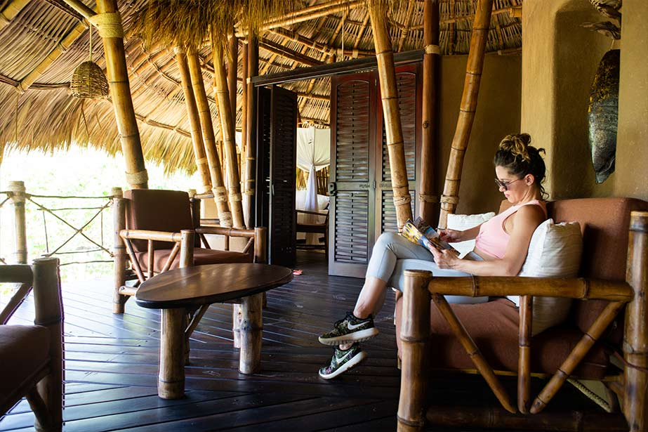 A person relaxes on a bamboo couch reading a book in an open-air, thatched-roof lounge overlooking a tropical area.