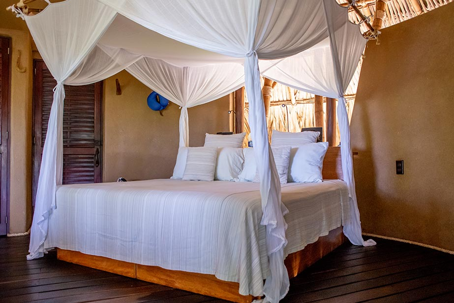 A bed with white linens and a canopy net inside a rustic room with earthy walls and wooden flooring.