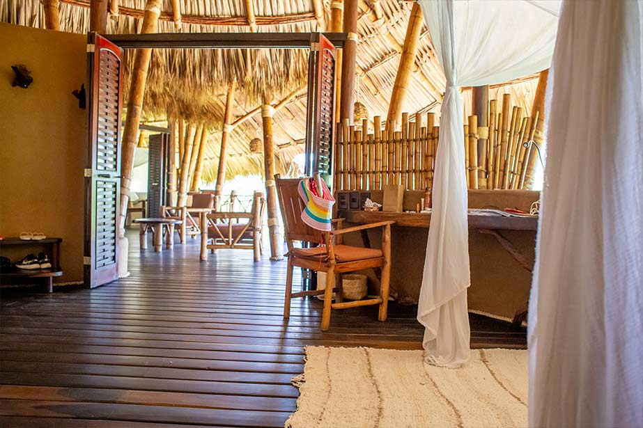 A wooden-floored lodge interior with a thatched roof, bamboo accents, a desk with a wooden chair, and hanging white curtains.