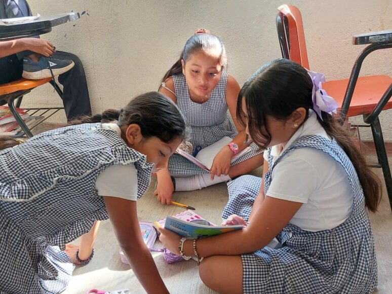 Three students in plaid uniforms sit on the floor around an open book, studying together indoors.