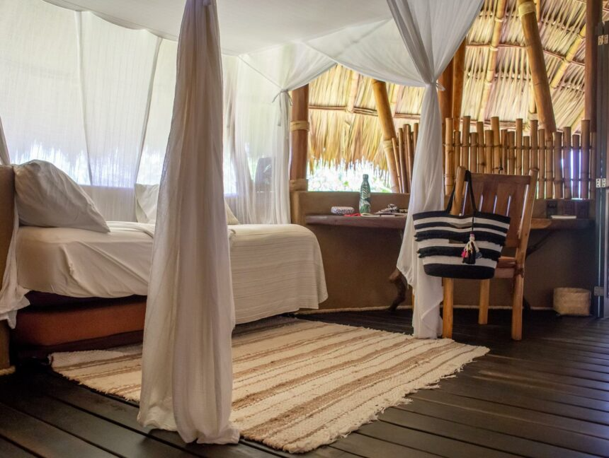 A canopy bed sits beside a woven rug, a wooden chair, and a striped tote bag in a rustic, thatch-roofed space.