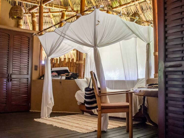 A canopy bed draped in white fabric sits in a rustic bedroom with wooden walls and a woven straw ceiling.
