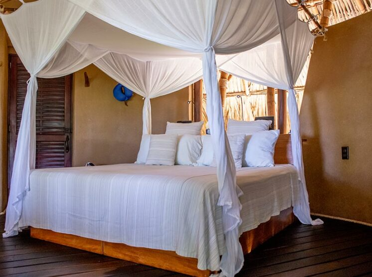 A canopy bed with white bedding in a rustic, earth-toned room with wooden floors and a thatched roof.