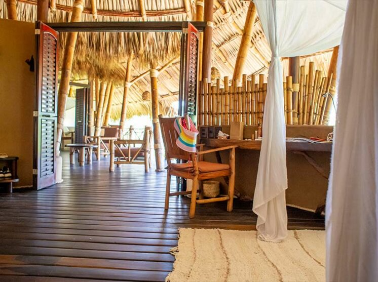 A rustic, open-air wooden room with a thatched roof, a chair with a colorful hat, a bamboo wall, and draped white curtains.