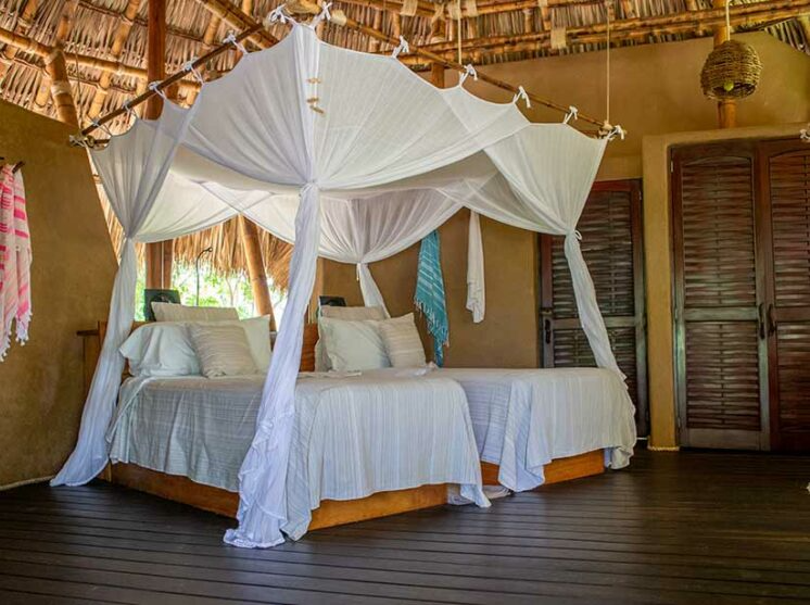 Two beds with white linens and draped mosquito netting in a rustic, thatched-roof room with wood flooring.