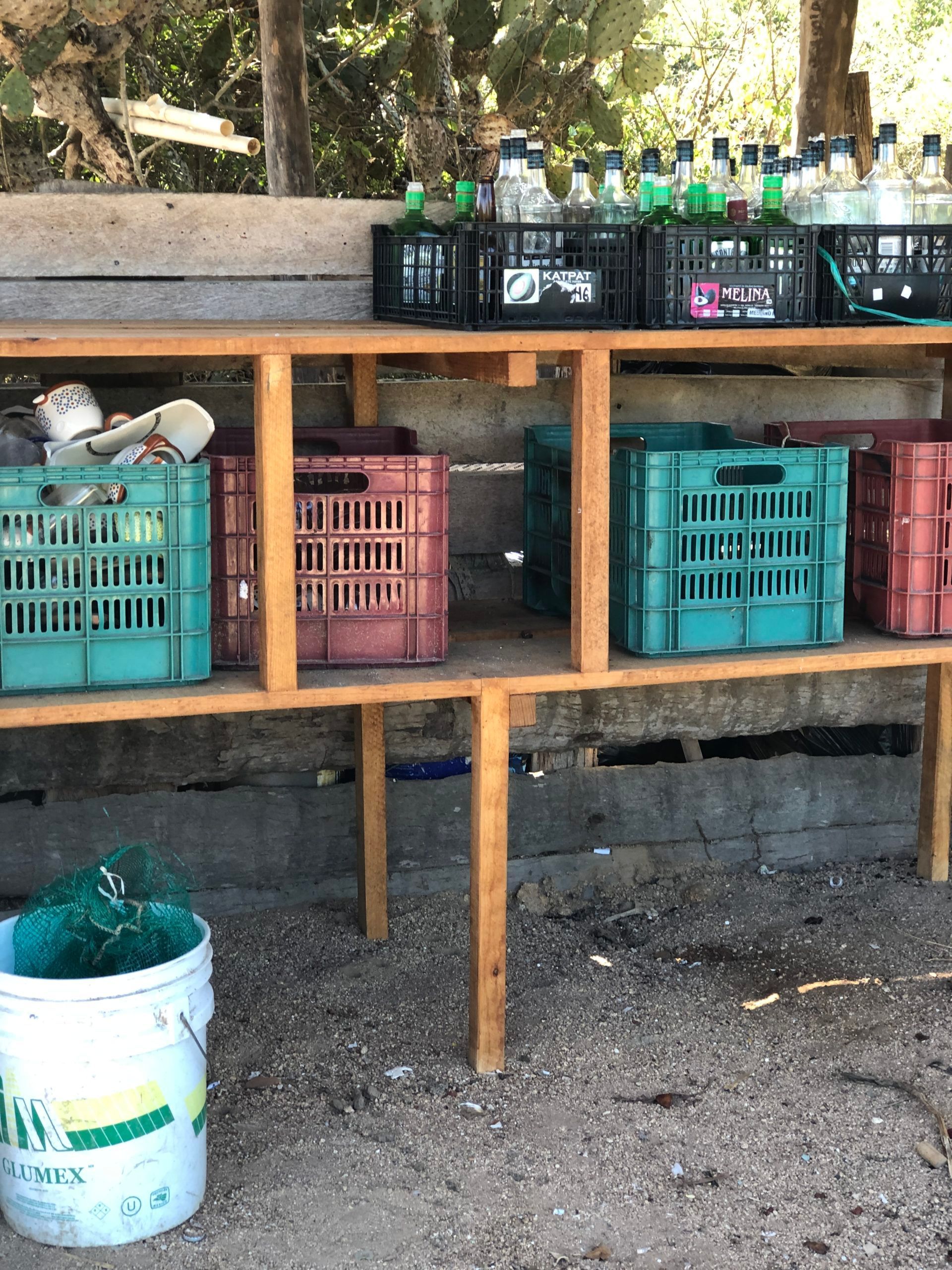 Outdoor wooden shelf with soda bottles and plastic crates beside a bucket on dirt ground