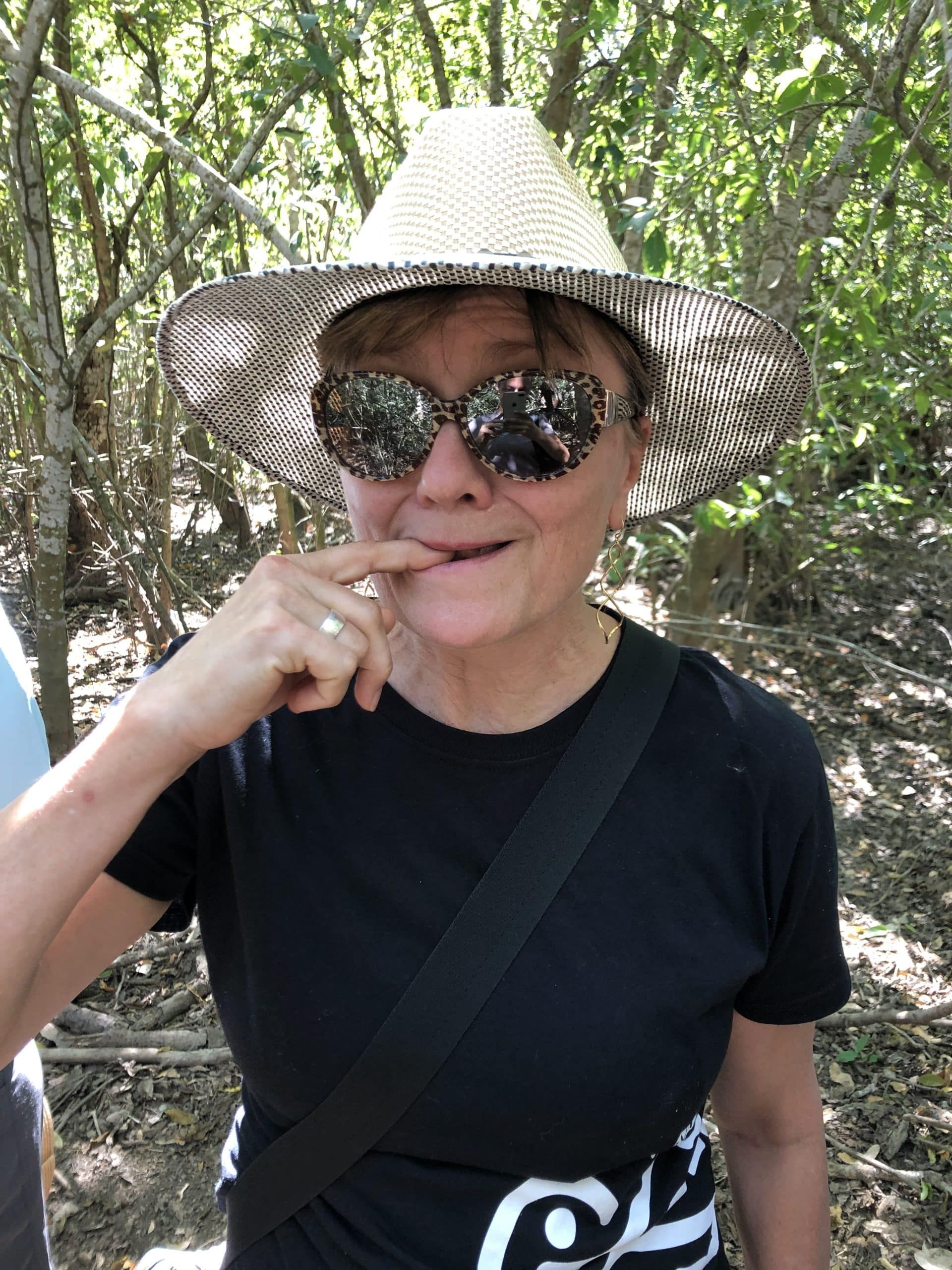 Person in a straw hat and sunglasses smiling outdoors among trees.