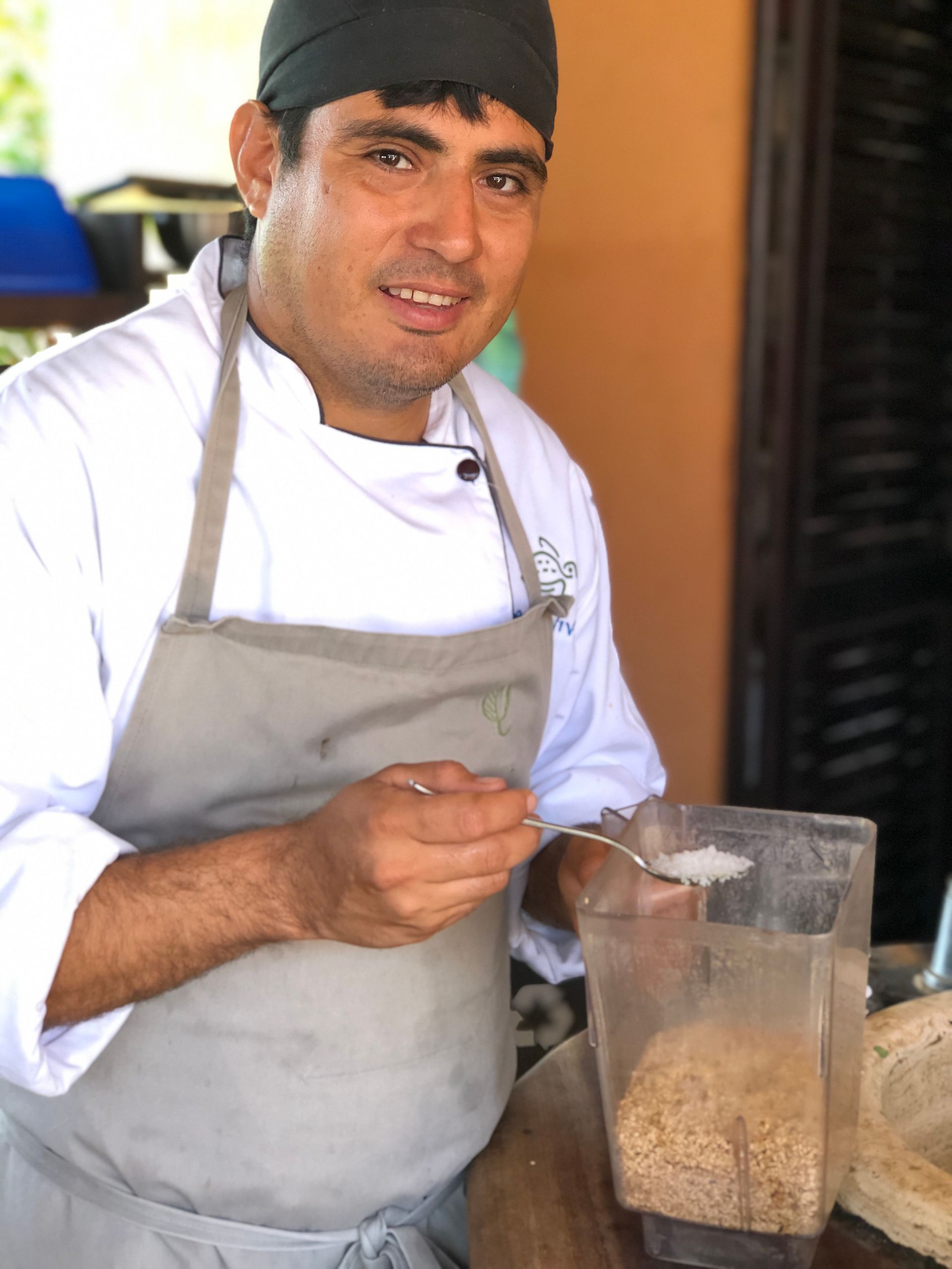Chef in white apron holding a spoon beside a blender filled with beans in a kitchen