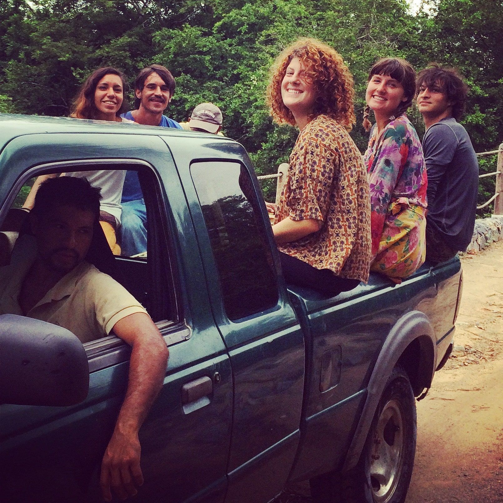 Five people riding in the back of a dark pickup truck on a dirt road, smiling outdoors.