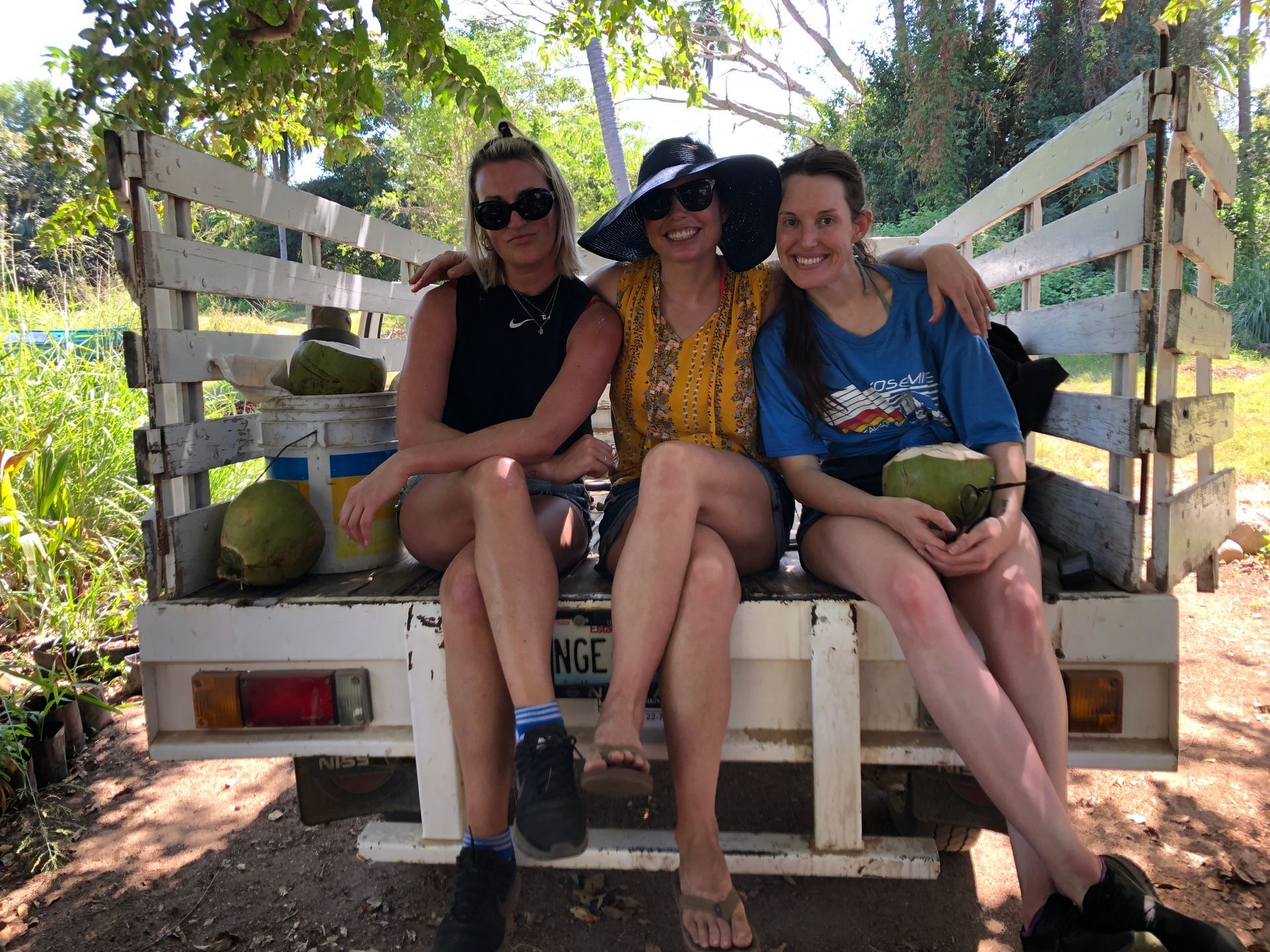 Three people sit on the back of a white truck in a sunny outdoor setting.