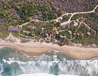 Aerial view of a coastal trail, sandy beach, and waves beside a lush seaside settlement