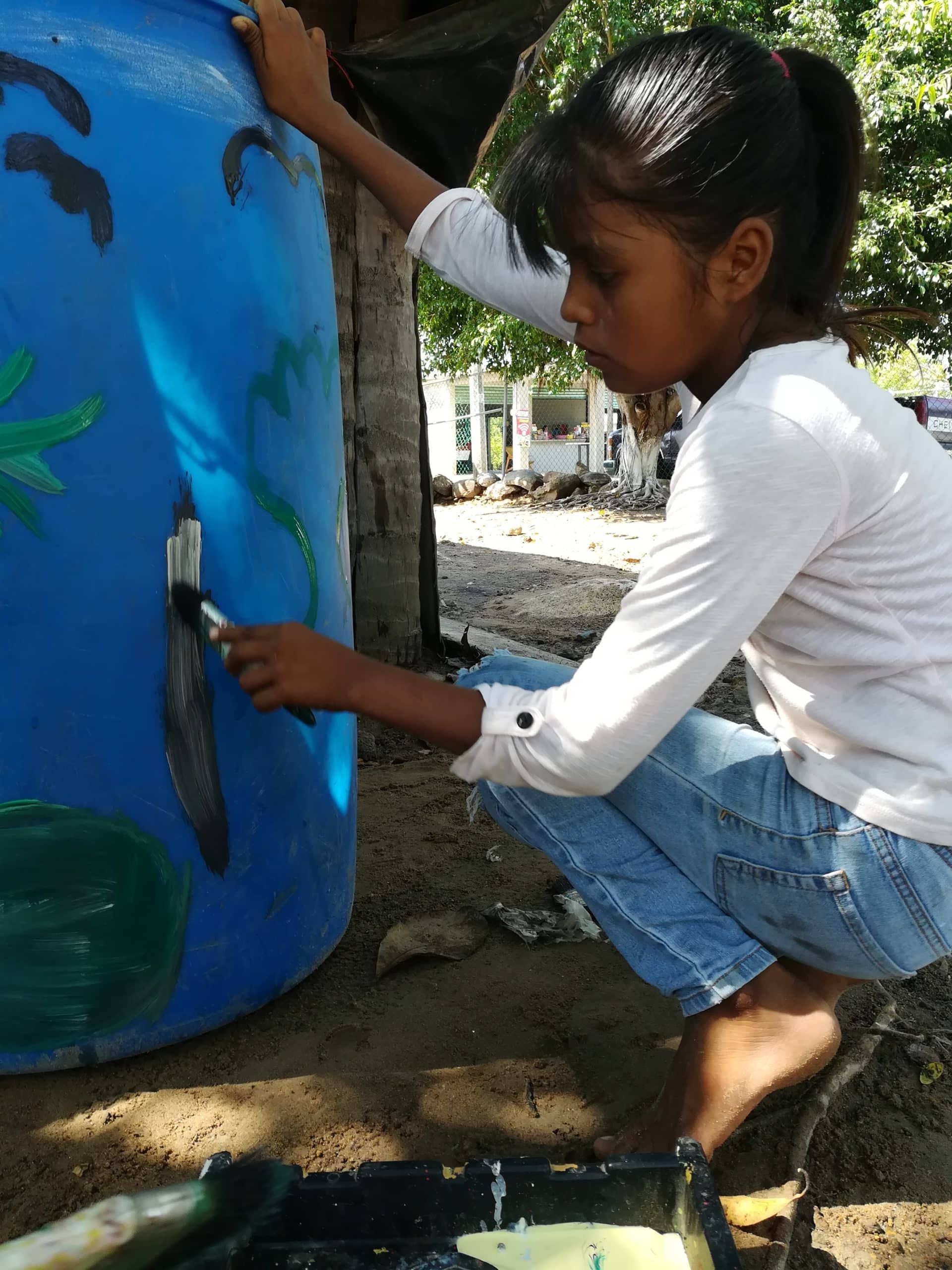 Person painting a large blue barrel outdoors with a brush, crouching beside it.