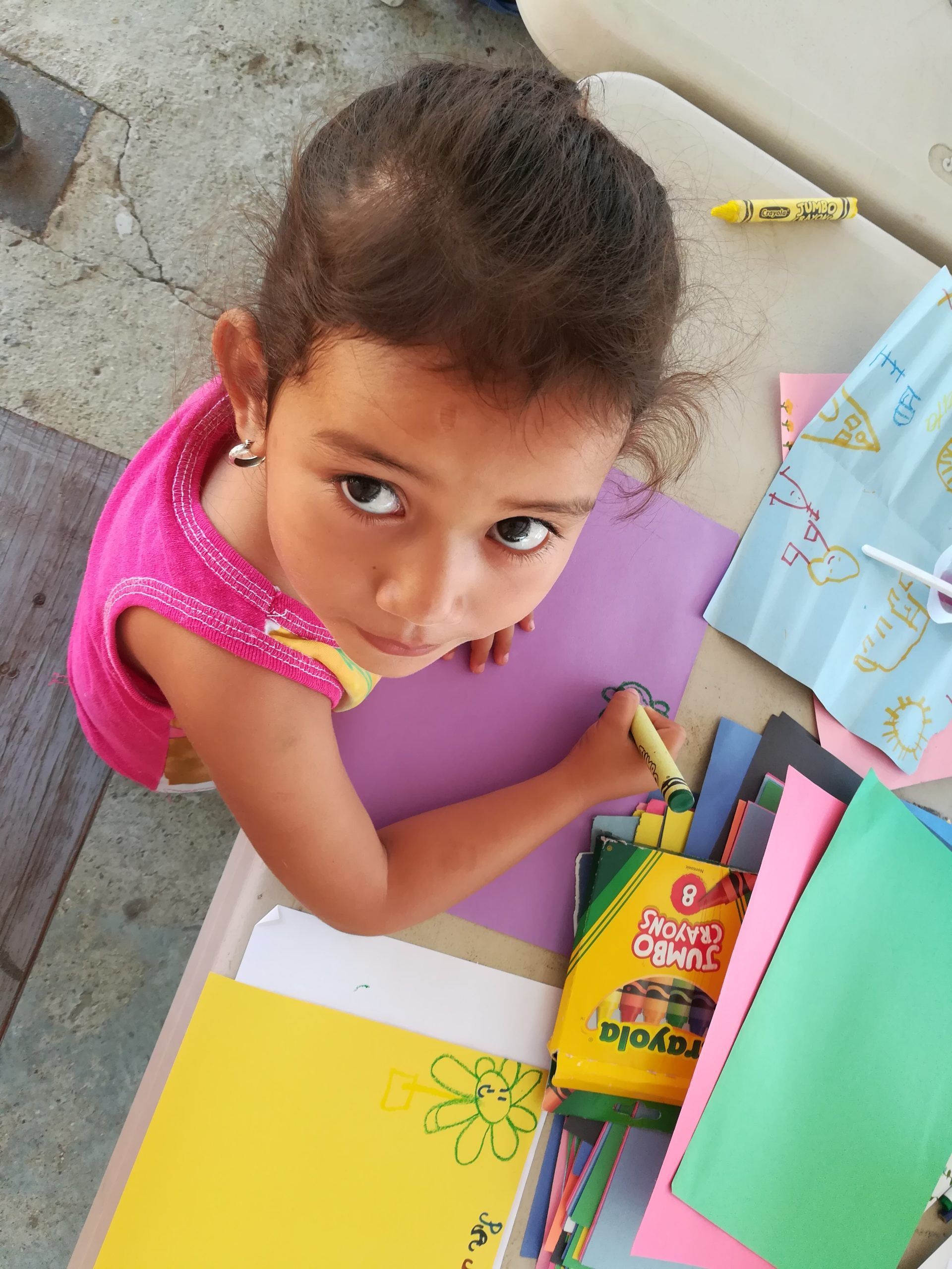 Child in a pink shirt looking up while drawing with crayons on a sidewalk.