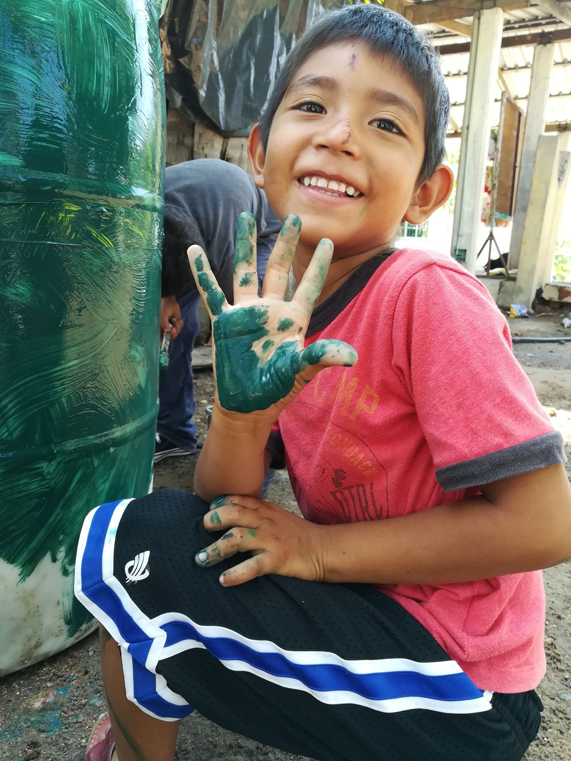 Smiling child showing a green-painted hand beside a large green barrel outdoors