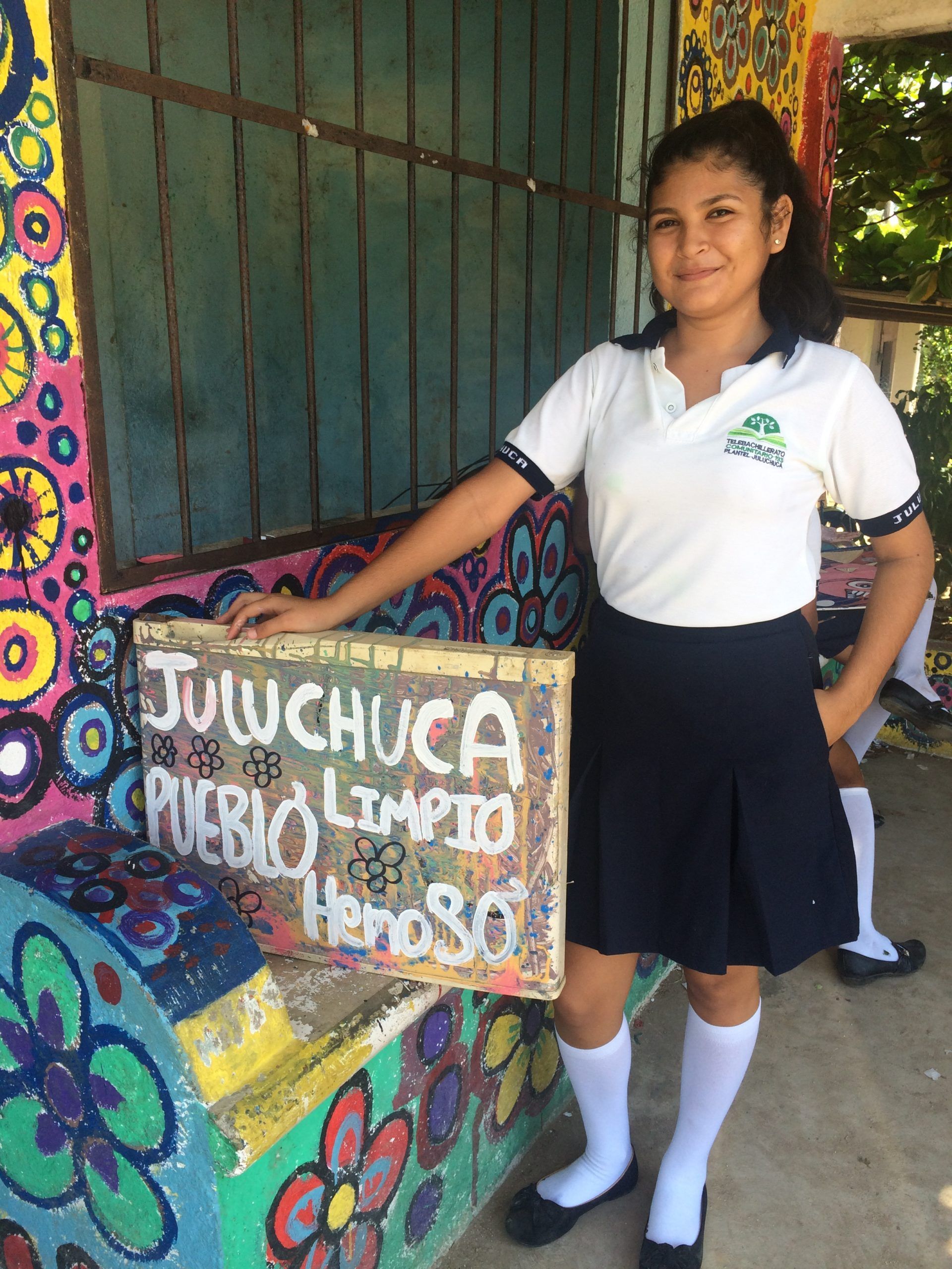 Student in white uniform stands beside a colorful mural and sign at a school entrance.