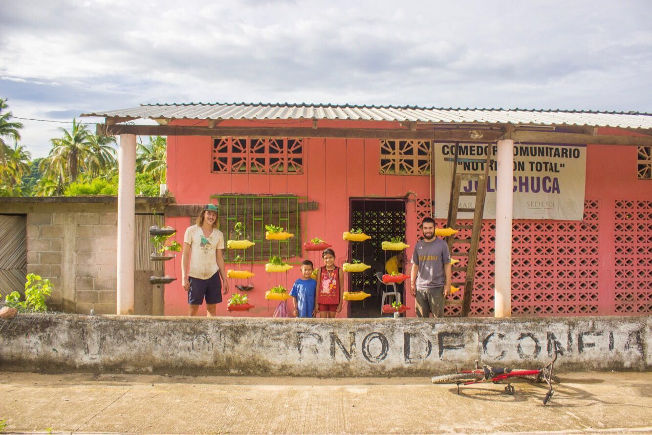 Small roadside shop with colorful goods and two people standing outside behind a low concrete wall.