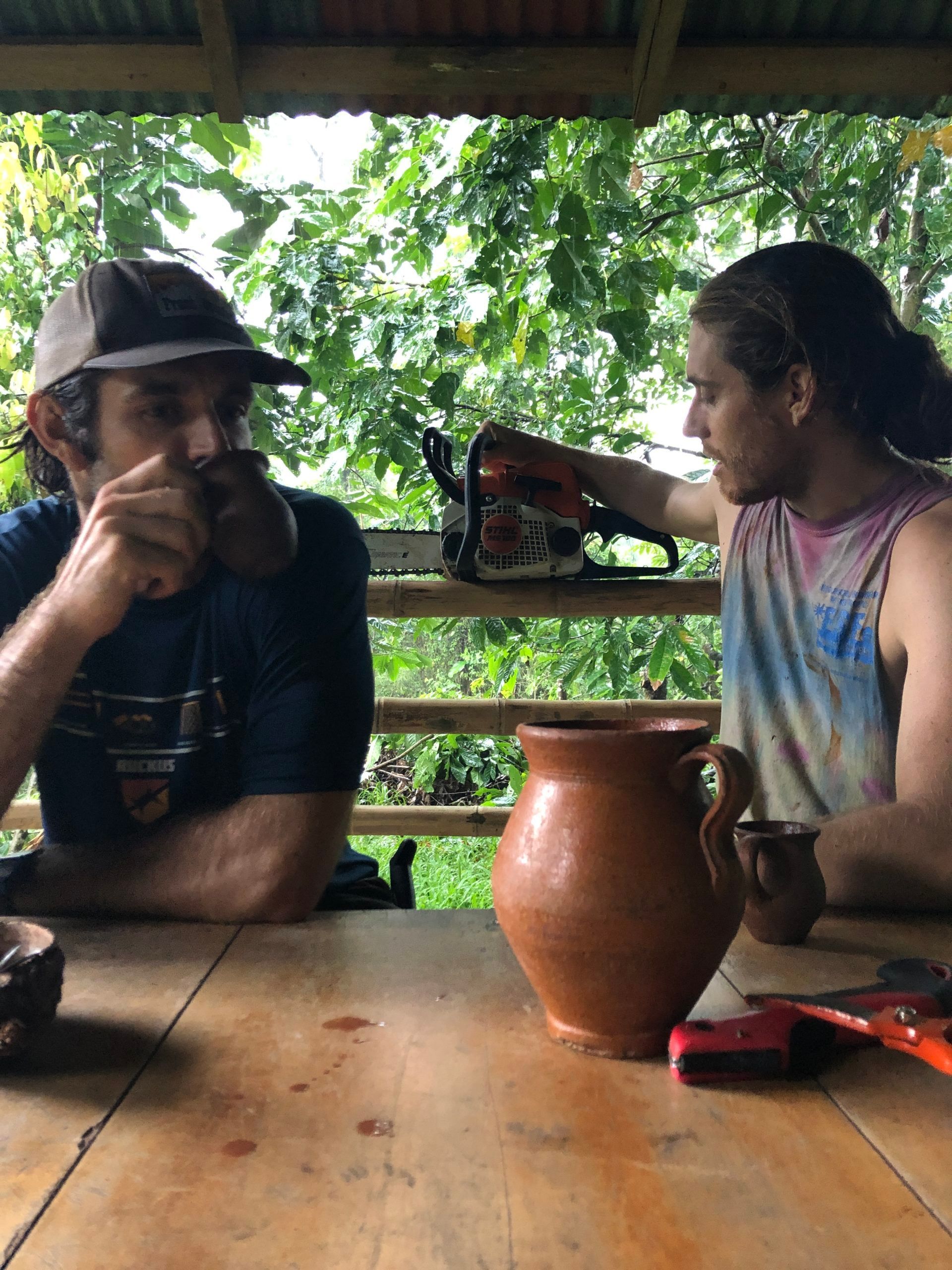 Two people sit at a wooden table under a porch, with a large clay pot and lush greenery behind them.