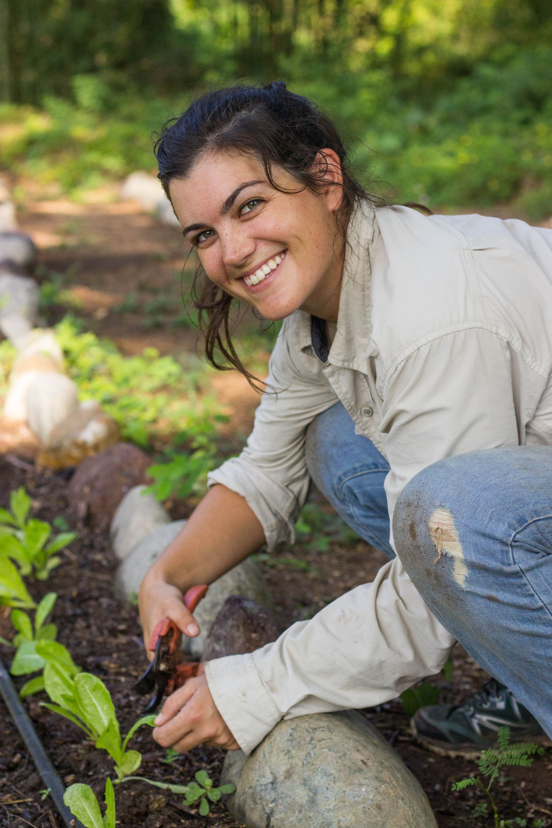 Person kneeling in a garden, planting seedlings in a raised bed and smiling at the camera.