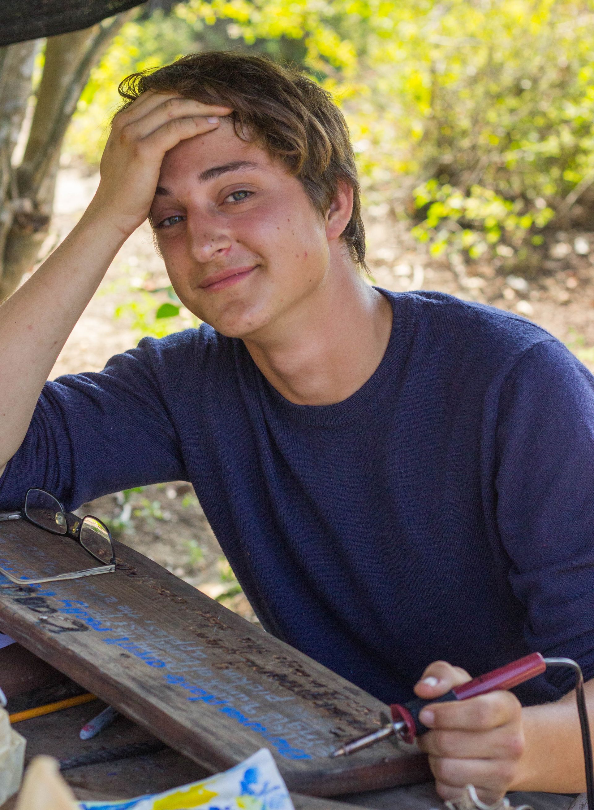 Person in a blue shirt smiling outdoors while holding their head beside a table with sunglasses.