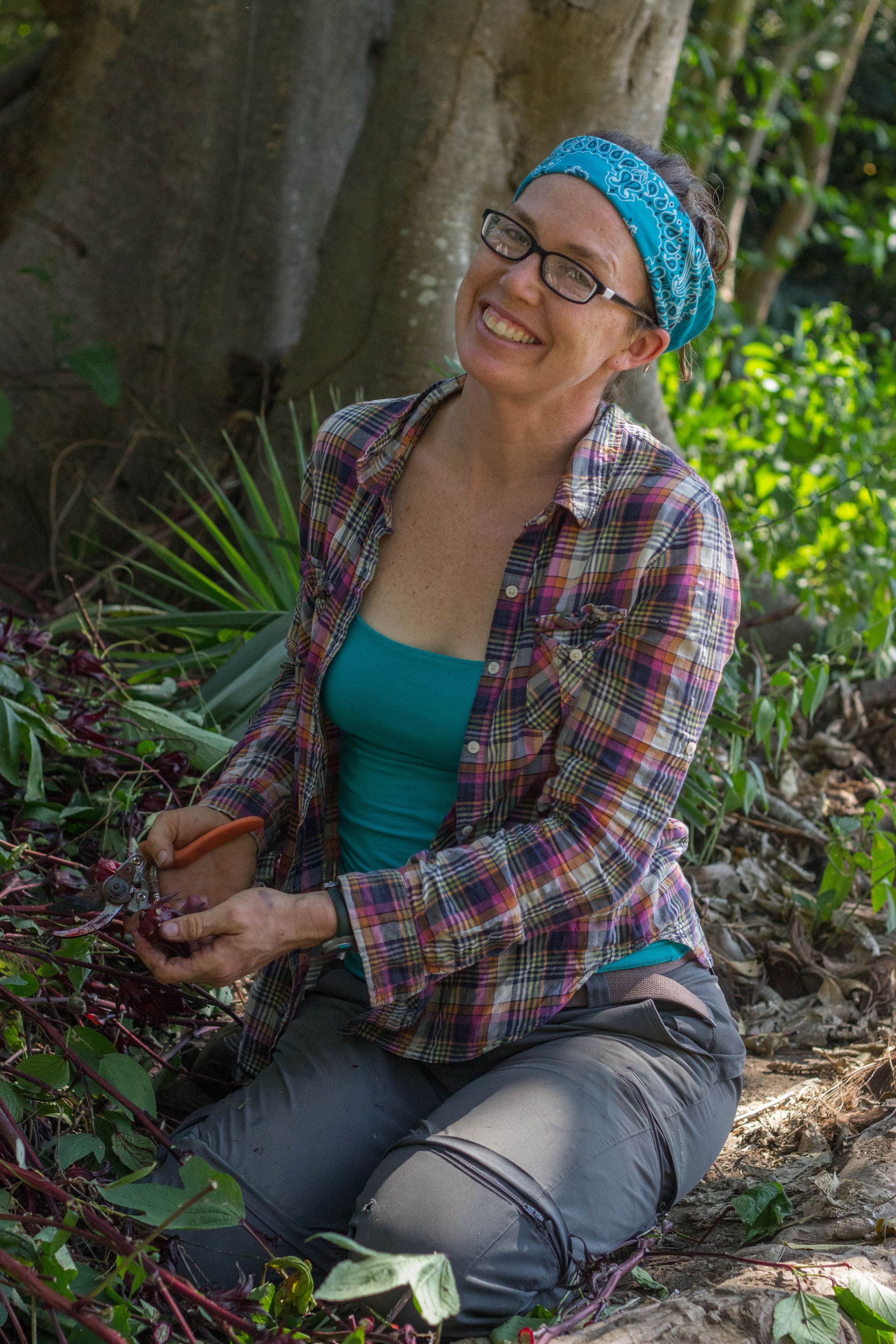 Smiling woman in a blue headband sitting outdoors by a tree, holding a small potted plant.