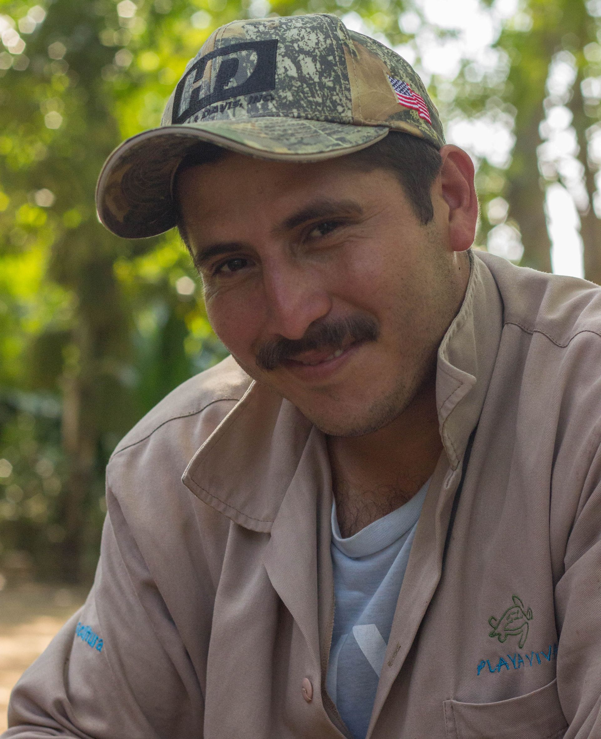 Man in a camouflage cap and tan shirt smiling outdoors among trees