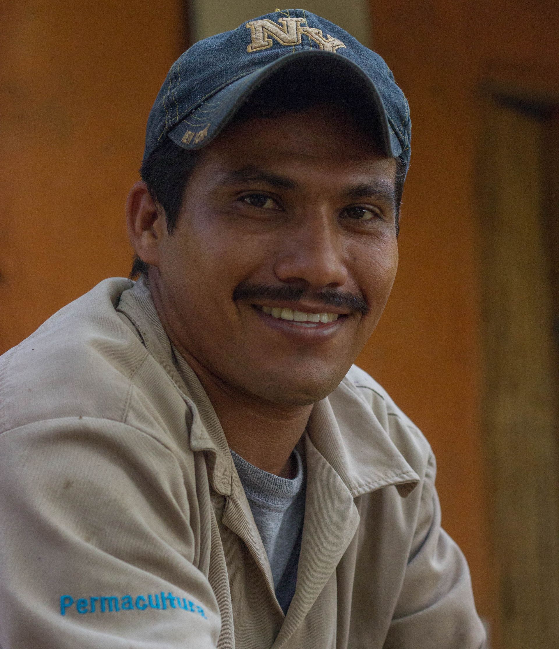Smiling man in a blue cap and beige jacket, seated against a warm brown background.