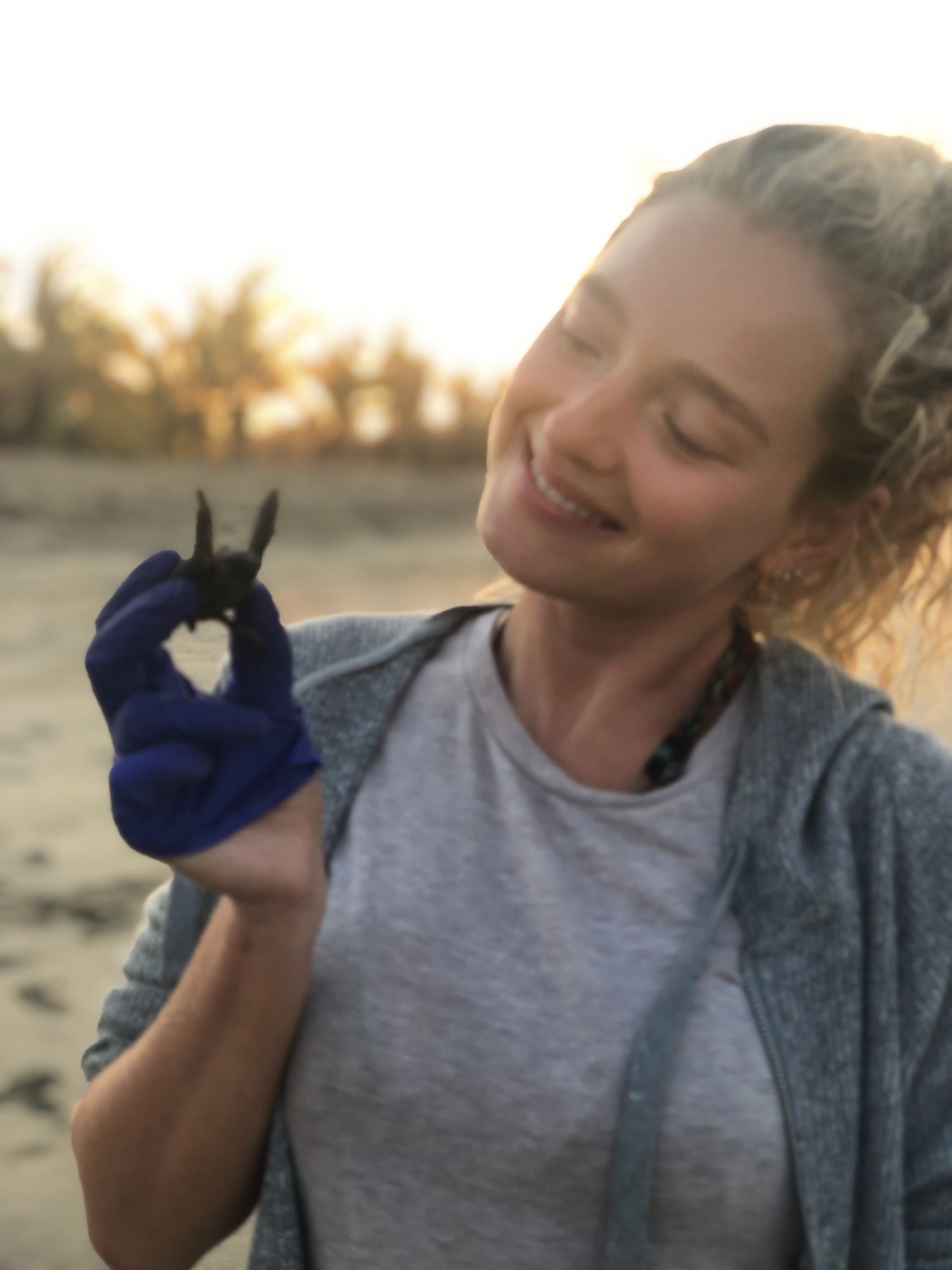 Woman smiling at a tiny black bird perched on her blue-gloved hand outdoors at sunset