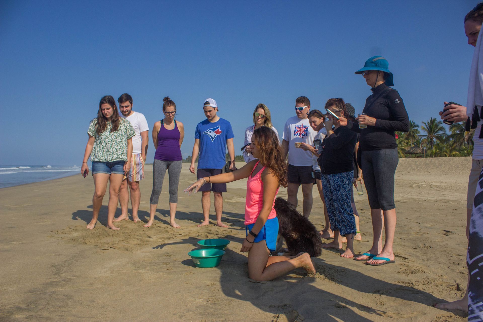 Group of people on a sandy beach, standing and kneeling near green buckets under a clear blue sky
