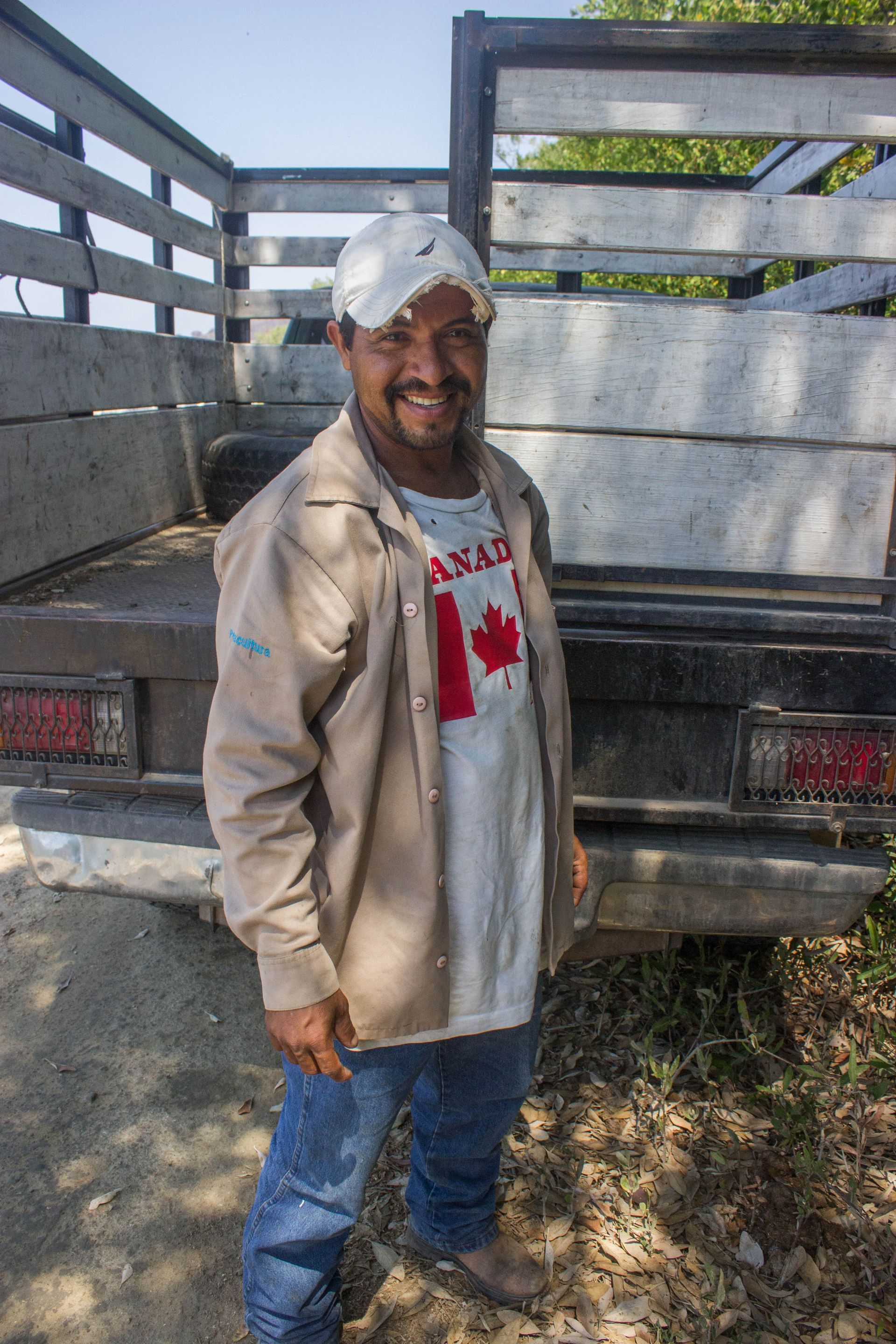 Man standing by a flatbed truck, wearing a cap, tan jacket, and jeans, smiling outdoors.