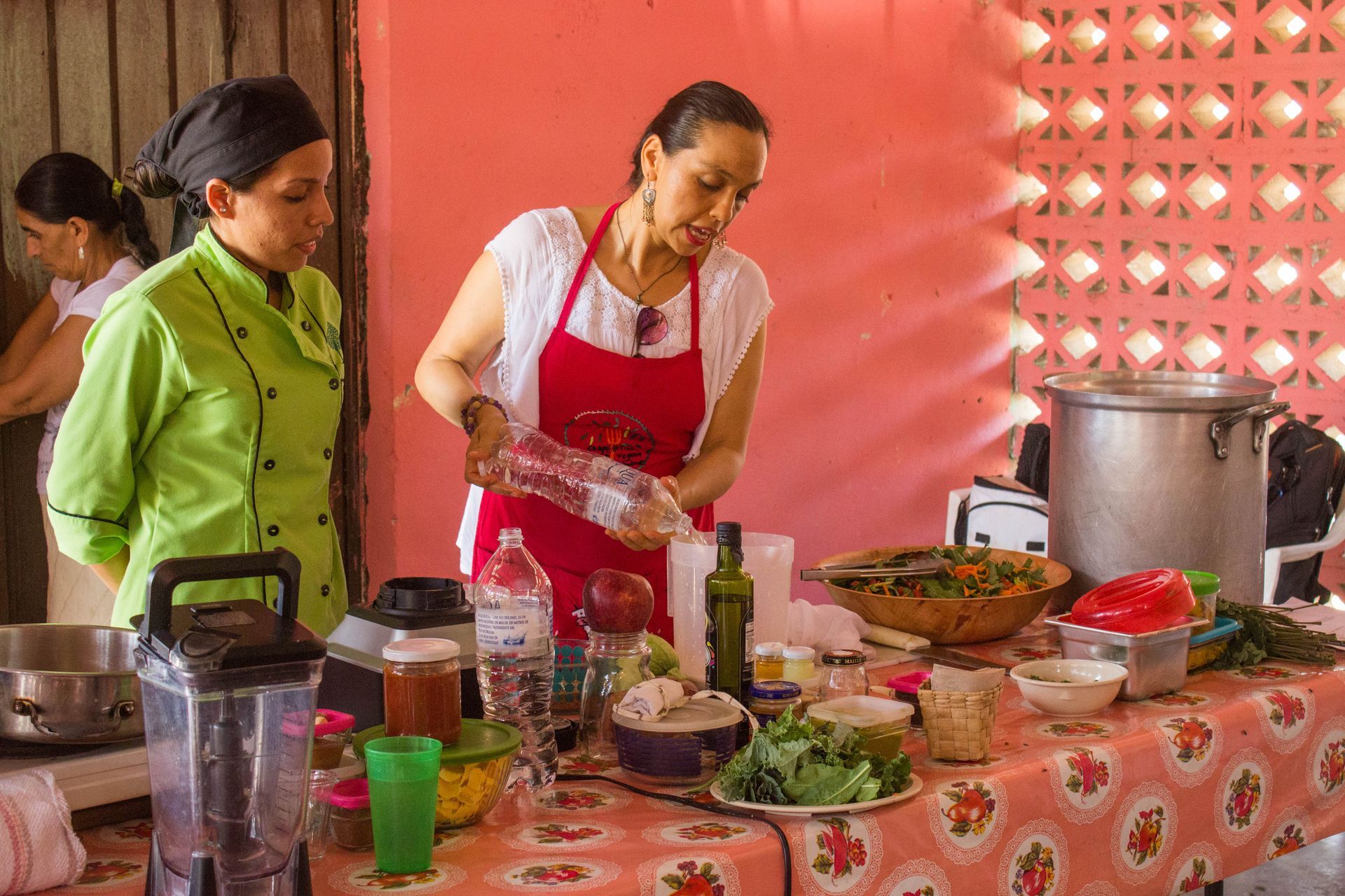 Two women prepare food at a buffet table with dishes, pots, and ingredients in a tented kitchen.