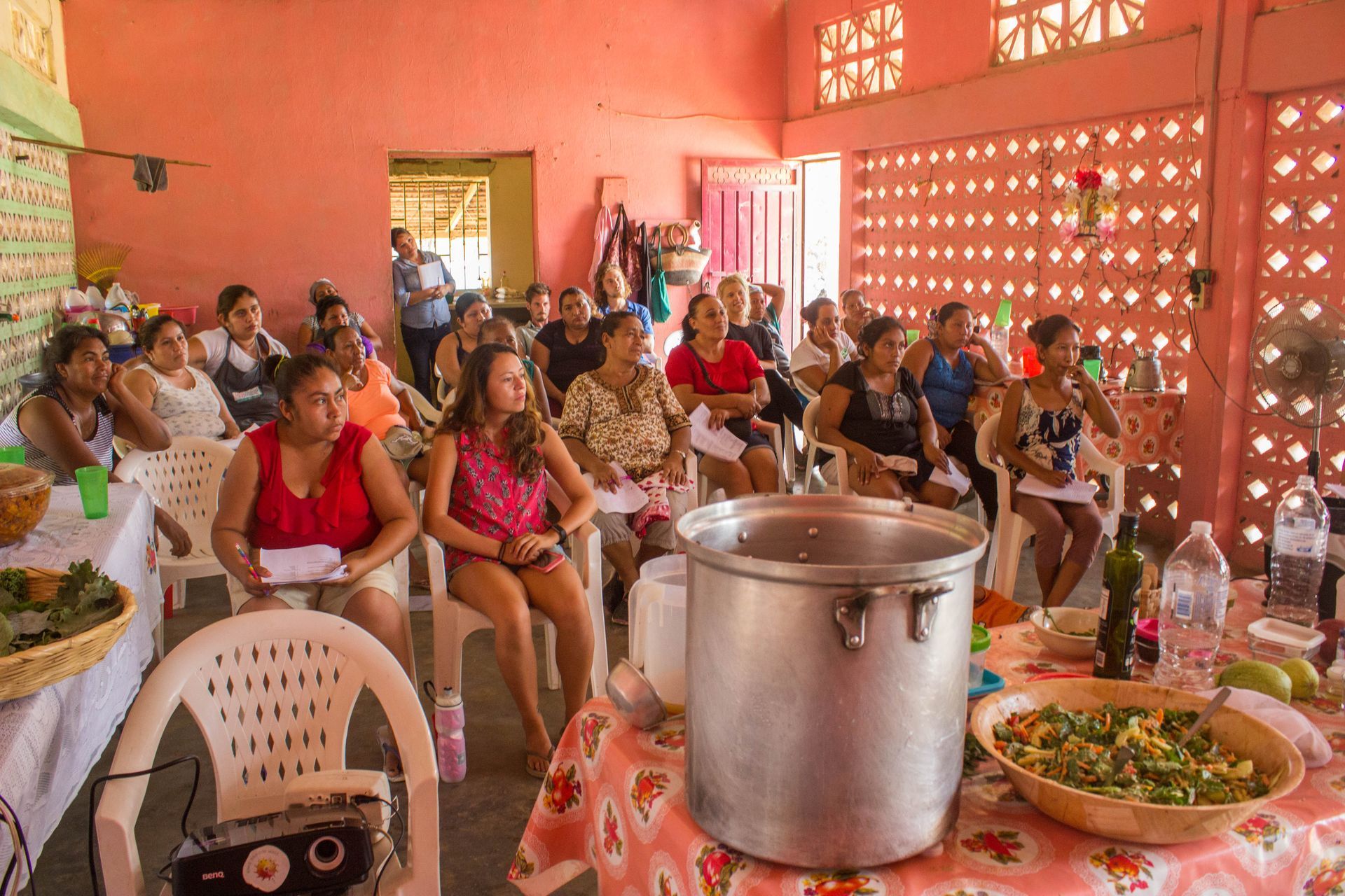 People seated in a pink room with food trays and a large metal pot on a table