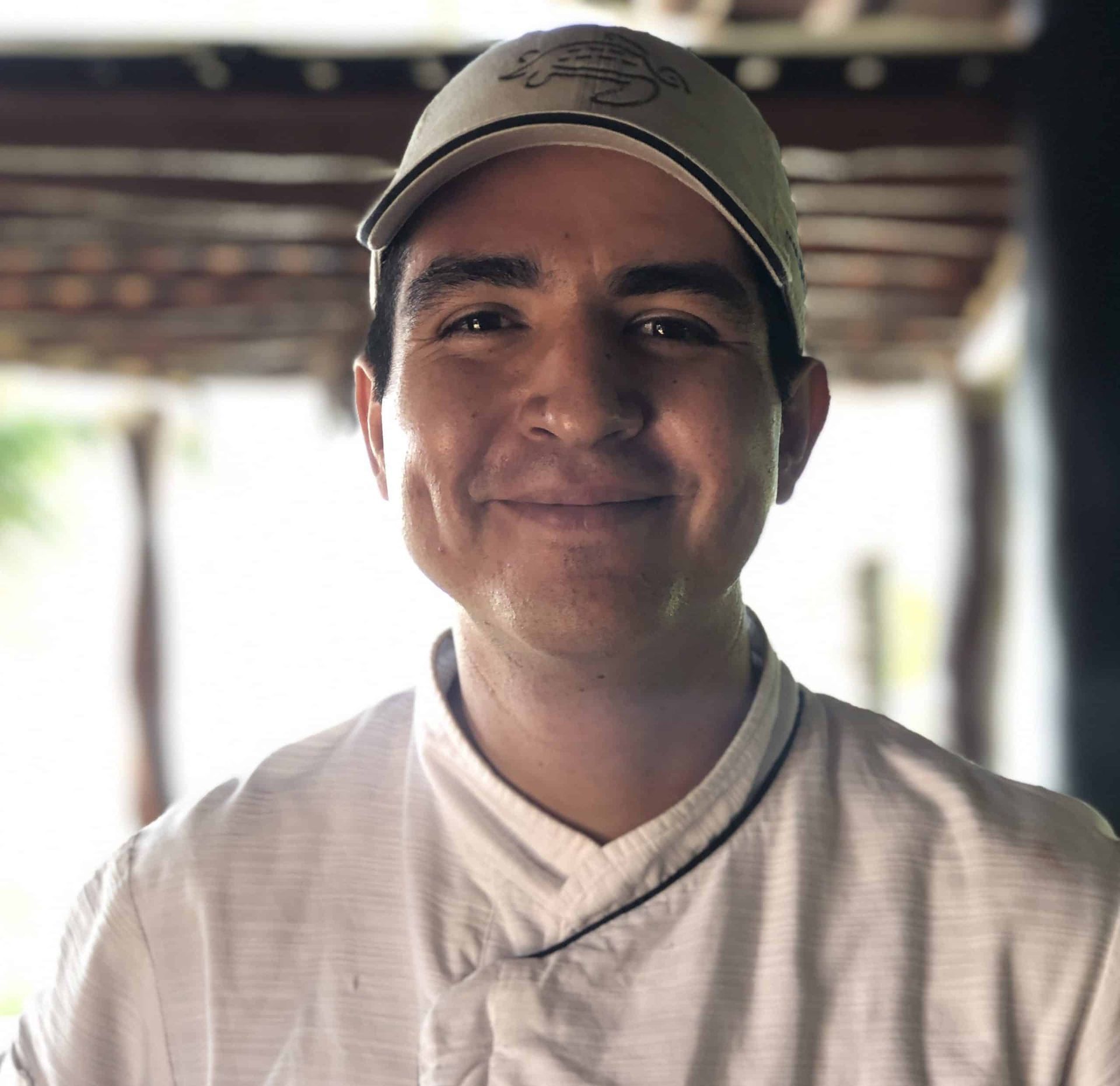 Smiling person in a light shirt and tan cap, standing indoors with a softly blurred background