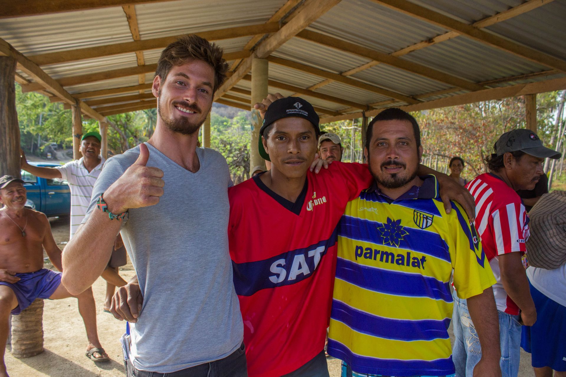 Three men posing together in a shaded outdoor shelter, smiling at the camera; one gives a thumbs-up.